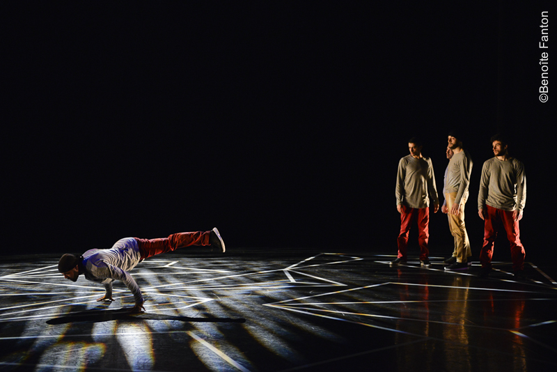 A dancer balancing on his hands on the floor, watched by three others standing, on a dark, light-streaked stage.