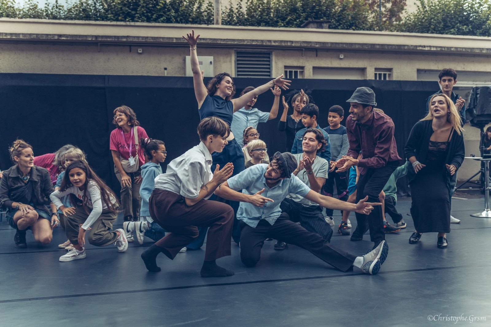 Atelier de danse en plein air : deux danseurs au sol en fente, entourés d'enfants et d'adultes qui lèvent les bras et applaudissent.